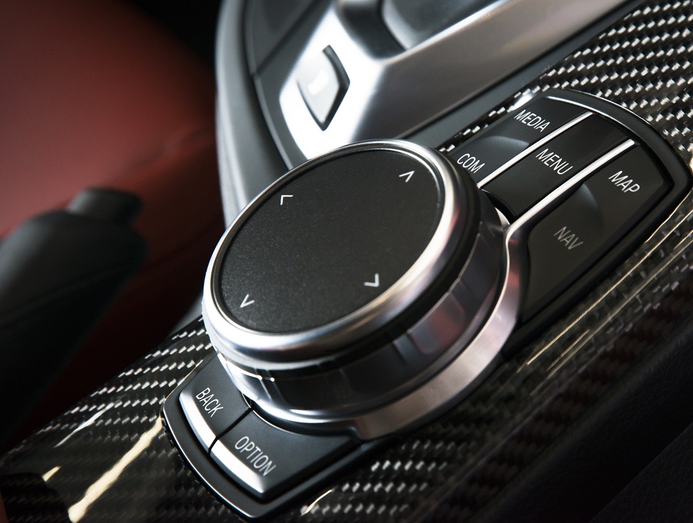 Close-up of a car's multimedia control dial and buttons—Media, Menu, Map, Nav, Com, Back, and Option—set in a carbon fiber-trimmed console, evoking the sleek precision found in top Audio and Lighting Services in New Orleans.