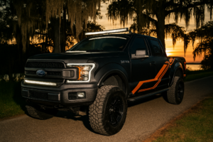 A matte black Ford F-150 with orange decals, a standout among custom builds Slidell, is parked on a road at sunset, surrounded by trees draped in Spanish moss.