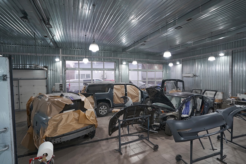 Large auto repair shop specializing in Restoration and Rebuilds in New Orleans, with several vehicles in various stages of repair; some cars have open hoods or covered parts, and car parts are visible around the workshop.