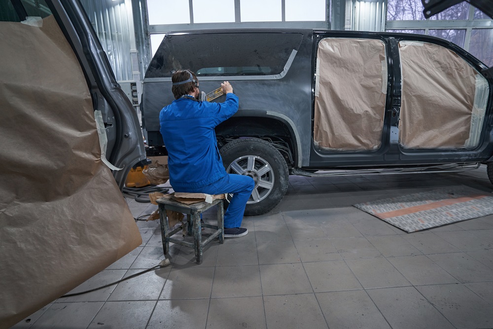 A person in blue coveralls repairs or paints the rear side of a car in an auto body shop, with paper masking the windows and doors—a scene common in Restoration and Rebuilds in New Orleans.